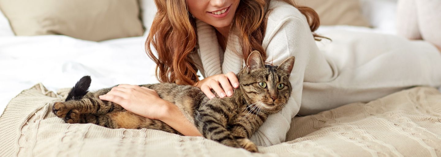 A woman in a cozy white sweater relaxes on a bed while gently petting a tabby cat lying beside her.